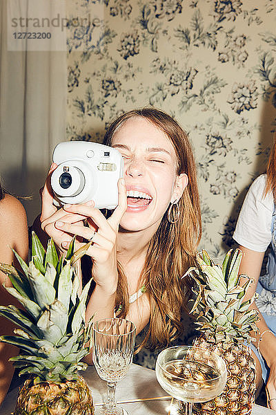 Cheerful young woman photographing while sitting amidst female friends at home during dinner party