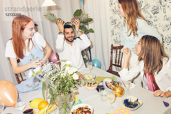 Playful man holding pineapples while sitting amidst female friends at home during dinner party