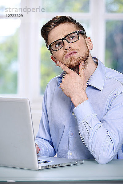 Mann in blauem Hemd  Brille  Bart  ernst  unscharfes Fenster im Hintergrund  sitzend  vor einem Laptop; Blick nach oben  2 Finger am Kinn  Blick nach oben.