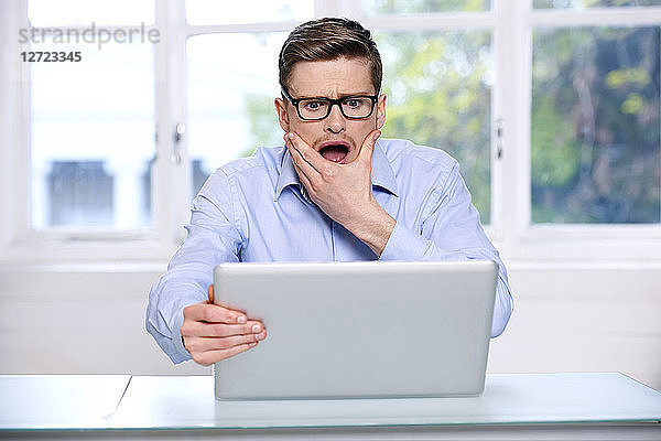 Mann in blauem Hemd; Brille; Bart; ernst; unscharfes Fenster im Hintergrund; geöffneter Mund; Hand im Gesicht; sitzend; Blick auf seinen Laptop; ängstlicher Blick