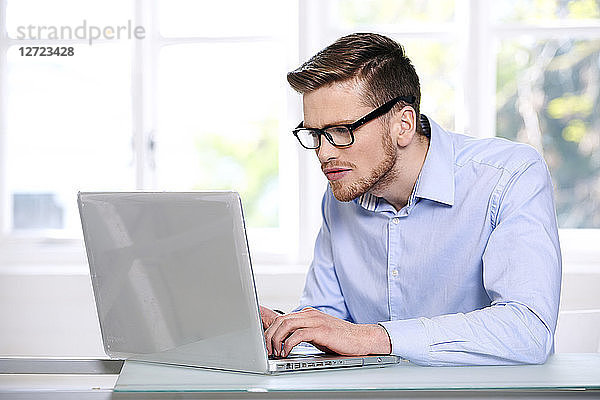 Mann im blauen Hemd  Brille  Bart  ernst  Fenster im Hintergrund unscharf  sitzend  tippt auf einem Computer-Laptop