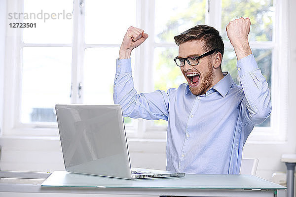 Mann mit blauem Hemd  Brille  Bart  lächelnd  unscharfes Fenster im Hintergrund  geöffneter Mund  Hände in der Luft  sitzend  auf seinen Laptop blickend .