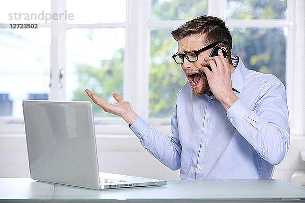 Mann in blauem Hemd; Brille; Bart; ernst; Fenster im Hintergrund unscharf; sitzend; Blick auf seinen Computer-Laptop; Telefon am Ohr  Hand in der Luft  schreiend .