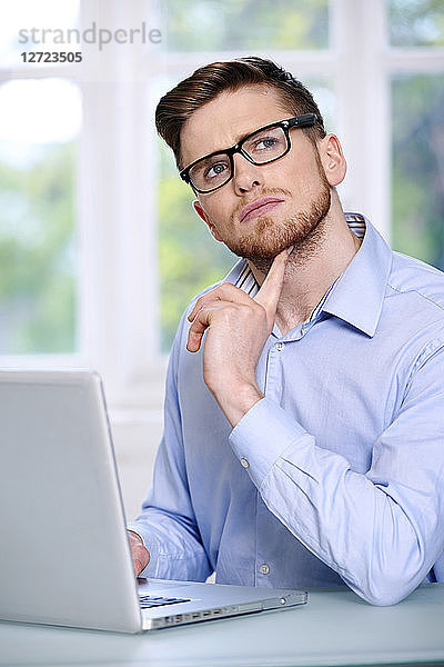Mann in blauem Hemd  Brille  Bart  ernst  unscharfes Fenster im Hintergrund  sitzend  vor einem Laptop; Blick nach oben  Finger unter dem Kinn  Blick nach oben.