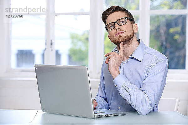Mann in blauem Hemd  Brille  Bart  ernst  Fenster unscharf im Hintergrund  sitzend  vor einem Laptop; aufblickend; nachdenklich; Finger am Kinn.