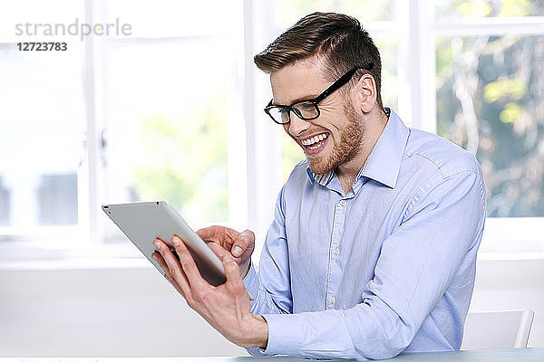 Mann in blauem Hemd; Brille; Bart; lächelnd; Fenster im Hintergrund unscharf; sitzend; schaut auf ein Tablet .