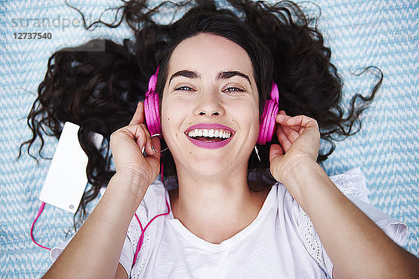 Portrait of happy young woman lying on blanket listening music with smartphone and pink headphones