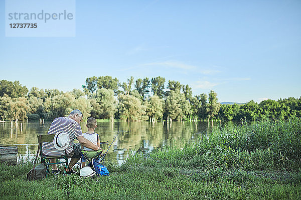 Back view of grandfather and grandson fishing together at lakeshore