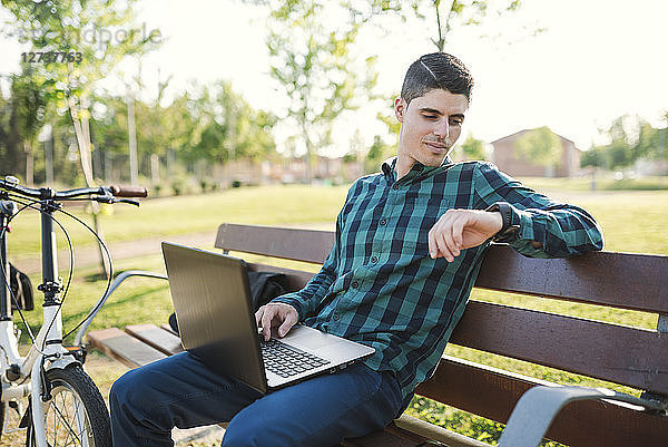 Young man with laptop on park bench checking the time