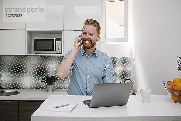 Businessman with laptop talking on the phone in his kitchen