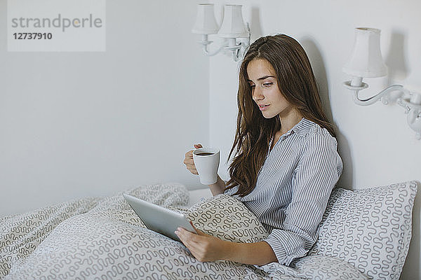 Woman using tablet while drinking coffee in bed in the morning