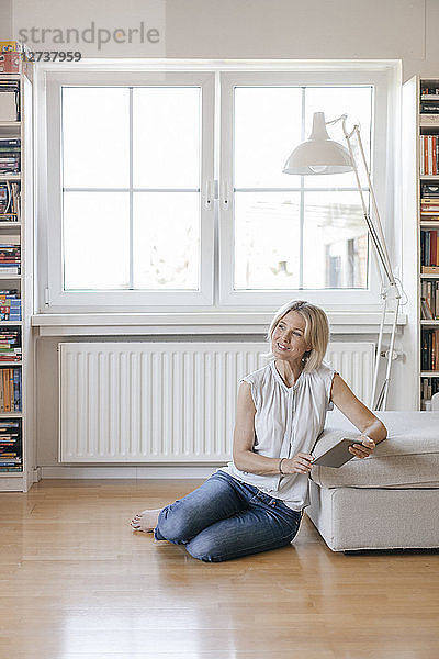 Smiling mature woman sitting on the floor at home holding tablet