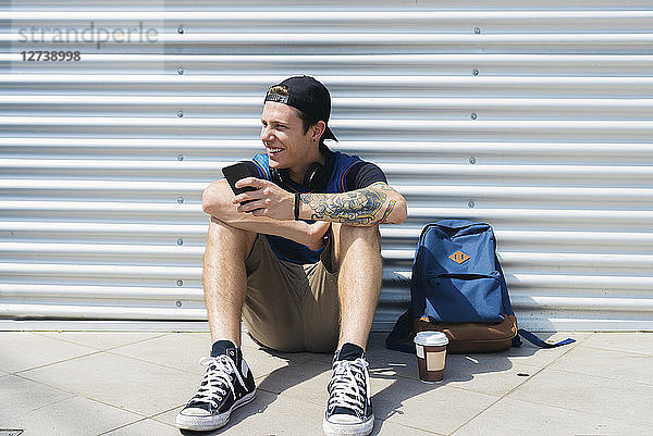 Smiling young man with smartphone  backpack and coffee to go sitting on the ground