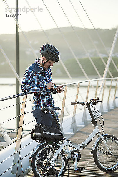 Young man with folding bicycle on a bridge looking at tablet