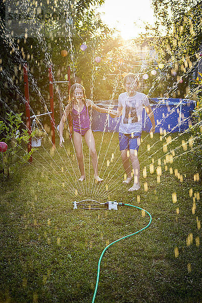 Brother and sister having fun with lawn sprinkler in the garden