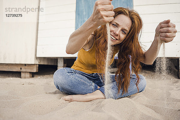 Redheaded woman sitting in front of beach cabin  with sand trickling through her hands