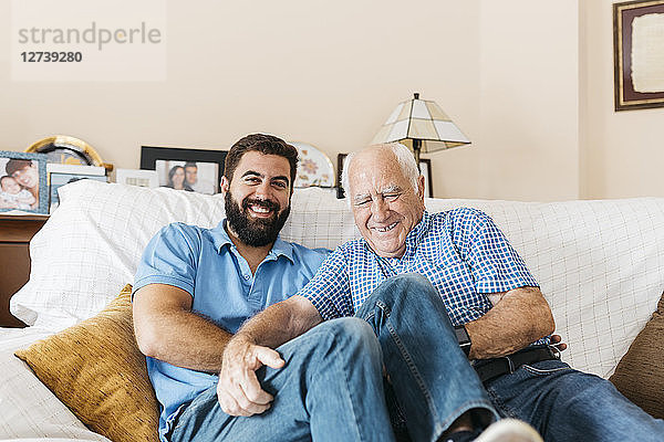 Portrait of adult grandson and his grandfather sitting on the couch at home tickling each other