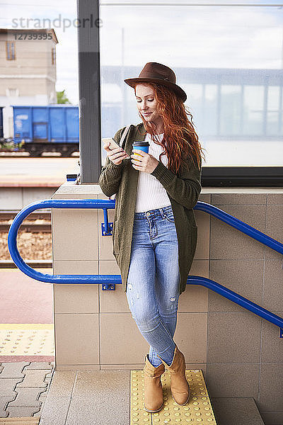 Smiling redheaded woman with coffee to go looking at cell phone at platform