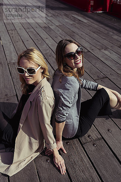 Portrait of two fashionable young women sitting on wooden floor wearing sunglasses