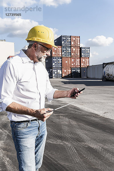 Businessman at cargo harbour  weraing safety helmet  using smartphone and digital tablet