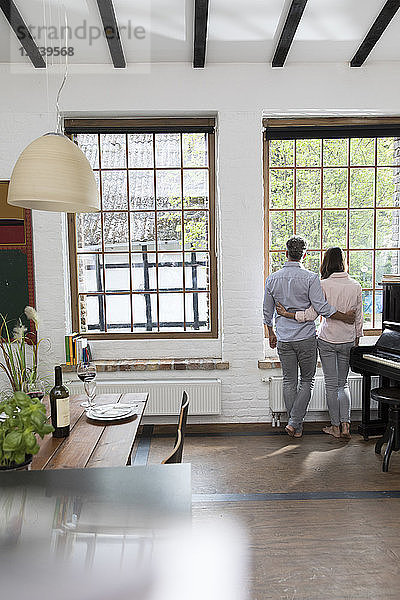 Mature couple standing in their comfortable loft  looking out of window  rear view