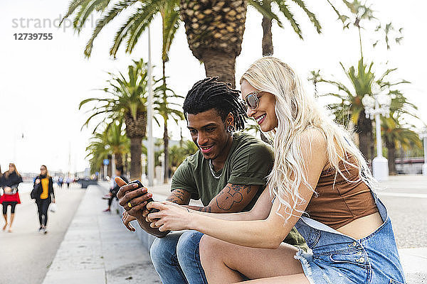 Spain  Barcelona  multicultural young couple sitting beside promenade looking at cell phone