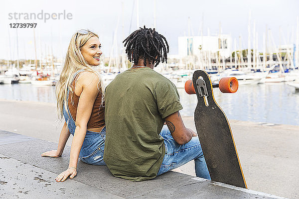 Spain  Barcelona  multicultural young couple with longboard sitting side by side at harbour