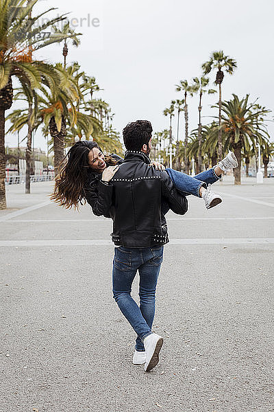 Spain  Barcelona  young man carrying happy girlfriend on promenade with palms