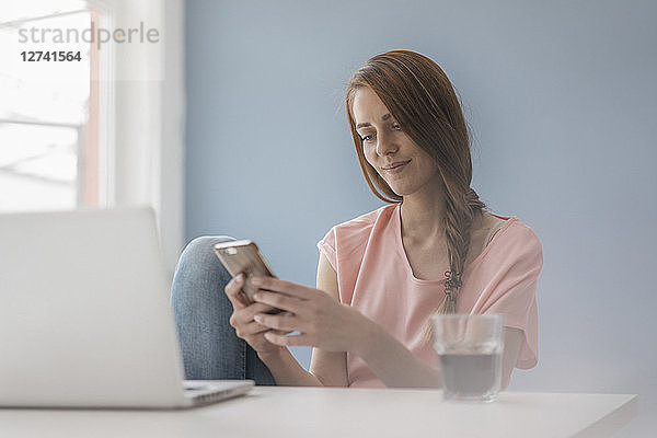Woman at home sitting at desk with laptop  using smartphone