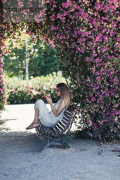 Woman with cell phone sitting on bench in park under pink blossoms