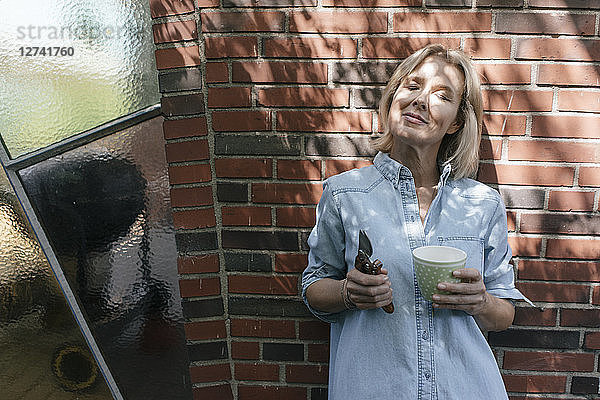 Relaxed mature woman leaning at house wall holding cup and pruner