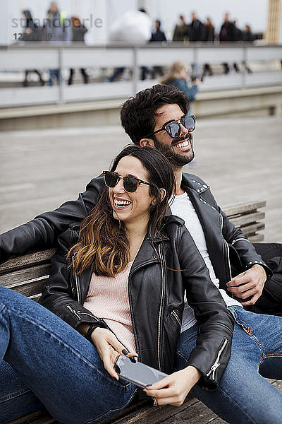 Spain  Barcelona  happy young couple with cell phone resting on a bench