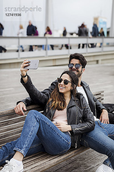 Spain  Barcelona  happy young couple resting on a bench taking a selfie