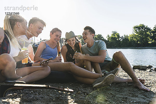Group of happy friends sitting at the riverside with drinks and cell phone