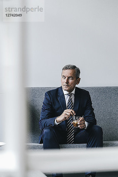Mature businessman with glass of coffee sitting on couch in his office looking out of window