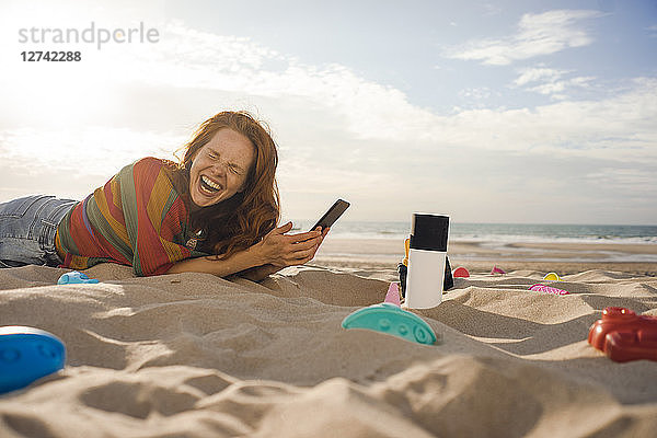 Redheaded woman lying on the beach with beach toys  using smartphone