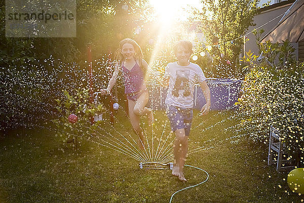 Brother and sister having fun with lawn sprinkler in the garden