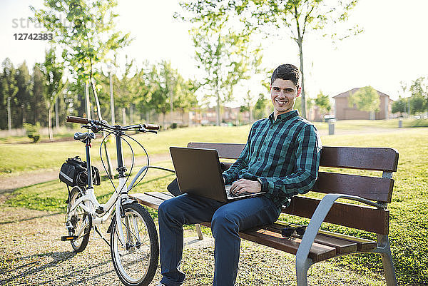 Young man with bicycle using laptop on park bench