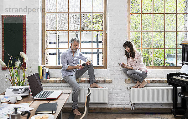 Mature couple working barefoot on window sill of home office  using smartphones