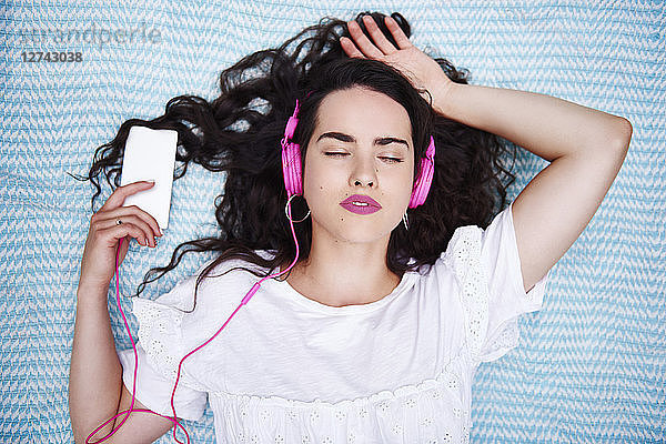 Portrait of young woman lying on blanket listening music with smartphone and pink headphones