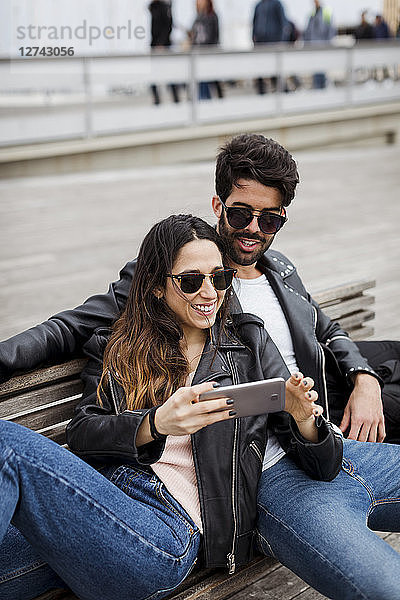Spain  Barcelona  happy young couple with cell phone resting on a bench