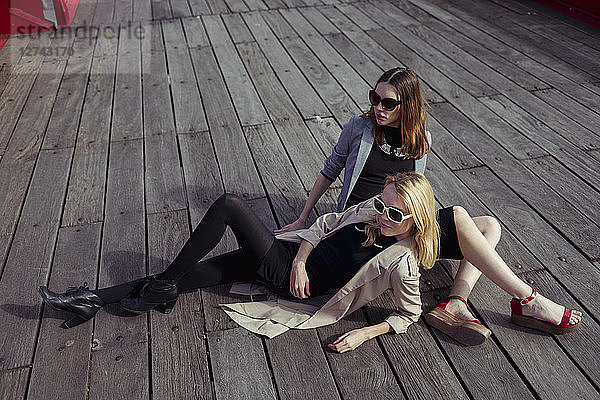 Two fashionable young women sitting on wooden floor wearing sunglasses