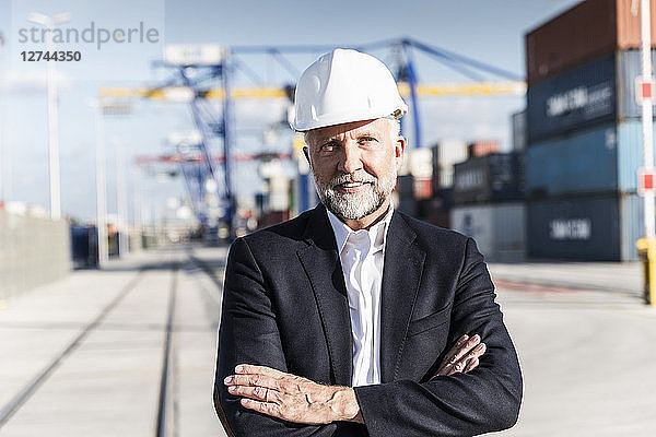 Businessman at cargo harbour  wearing safety helmet  arms crossed