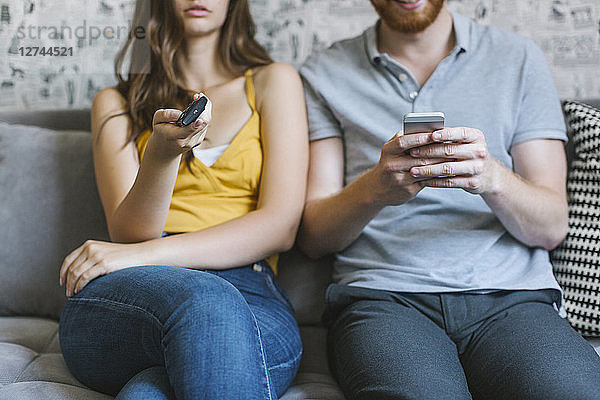 Couple sitting on couch with remote control and smartphone  partial view