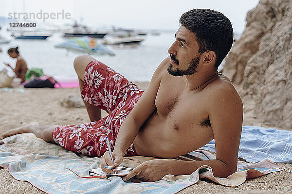Young man lying on towel at the beach  notebook  thoughtful