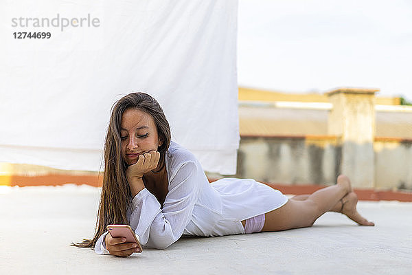 Portrait of smiling young woman lying on roof terrace looking at cell phone