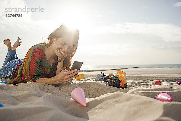Redheaded woman lying on the beach with beach toys  using smartphone