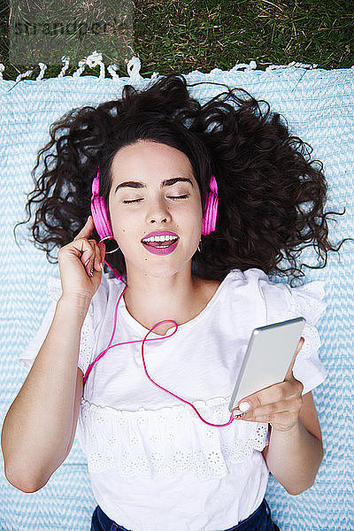 Portrait of young woman lying on blanket listening music with smartphone and pink headphones