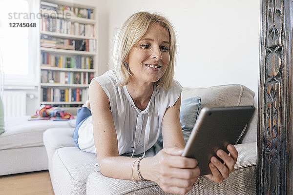 Smiling mature woman lying on couch at home using tablet