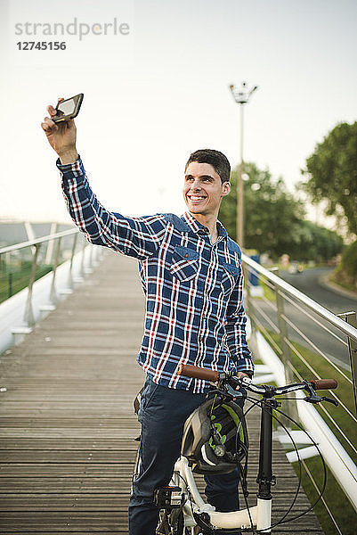 Smiling young man with folding bicycle on a bridge taking a selfie